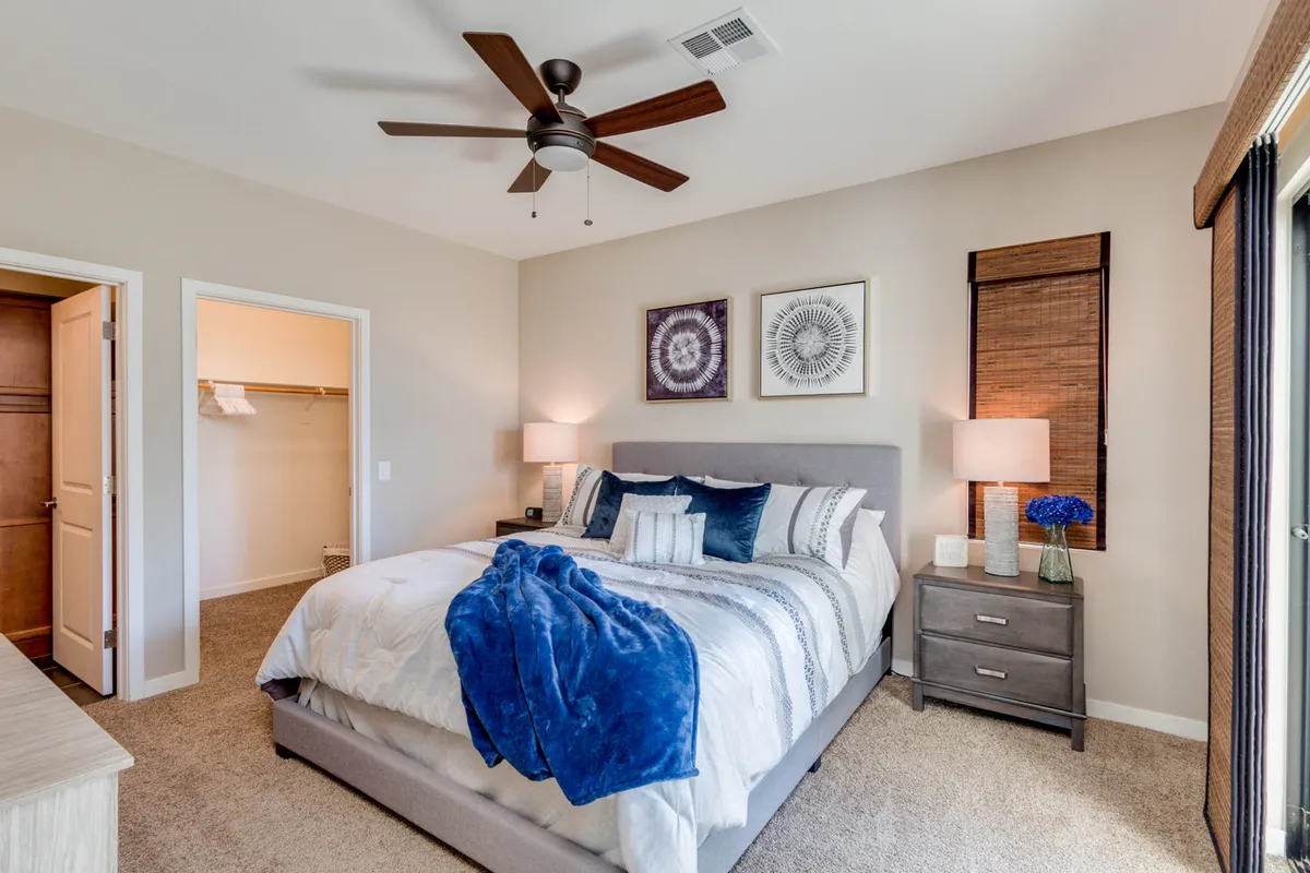 A spacious king-size bed dressed in soft, luxurious linens and decorative pillows sits against a calming neutral backdrop. A bamboo window treatment filters natural light softly into the room, and understated artwork on the wall adds a designer touch. A nightstand with a lamp provides a cozy reading nook, further enhancing the relaxing mood of the suite.