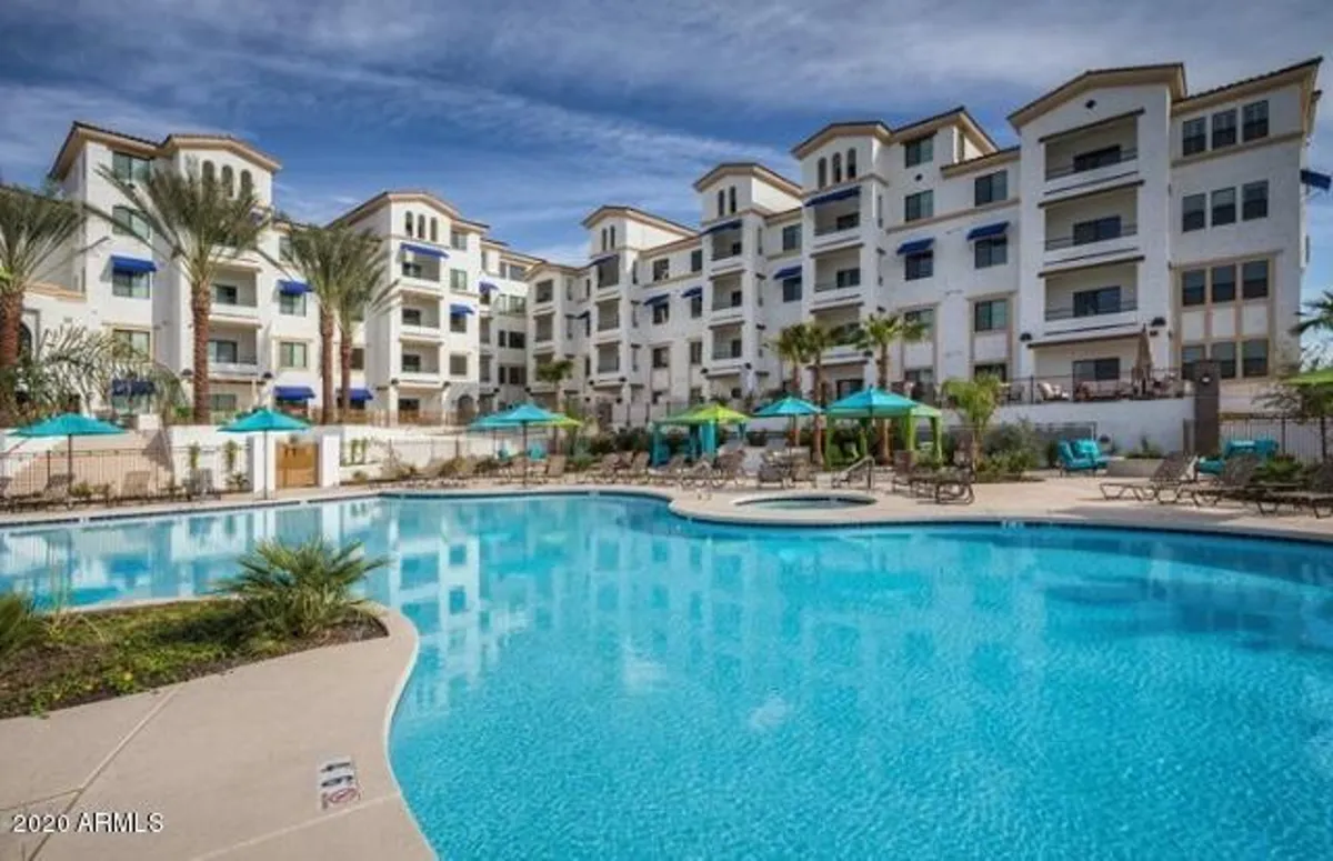 A sunlit exterior pool scene featuring a sparkling swimming pool, lounge chairs, umbrellas, palm trees, and the adjacent residential building. The setting is perfect for sunbathing, socializing, and enjoying meals outdoors.