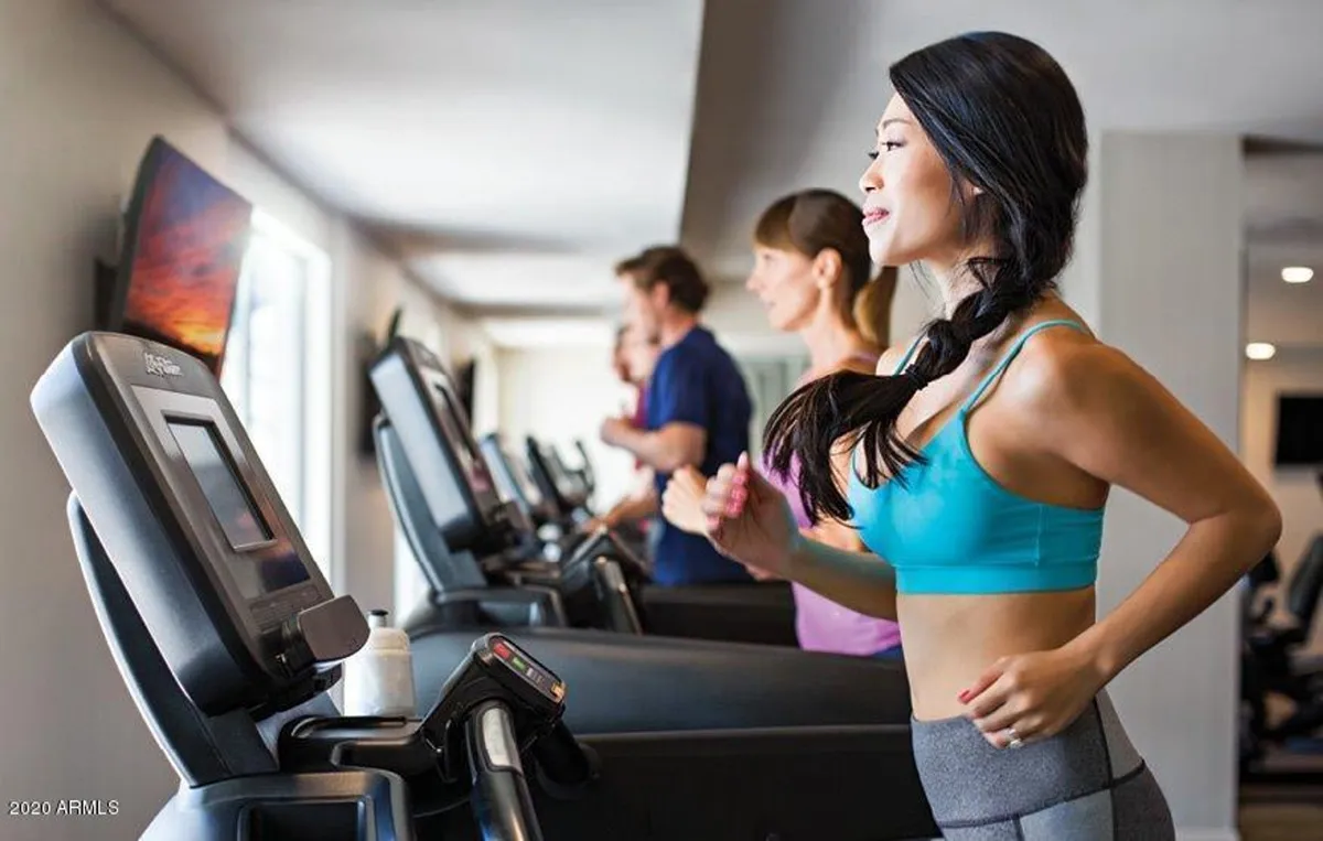 A modern fitness room featuring multiple treadmills, people actively exercising, and a wall-mounted TV. The environment is bright and motivational, fostering both wellness and social interaction.