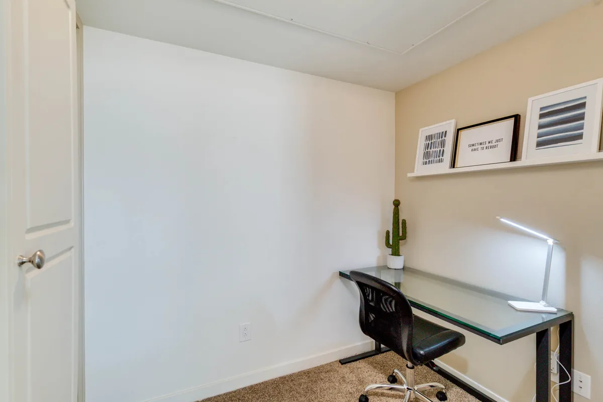 A minimalist office nook featuring a glass desk, black chair, cactus plant, and shelves with framed art, set in a quiet corner designed for productivity. The clean, organized setup offers a dedicated space for remote work or creative pursuits within the great room.
