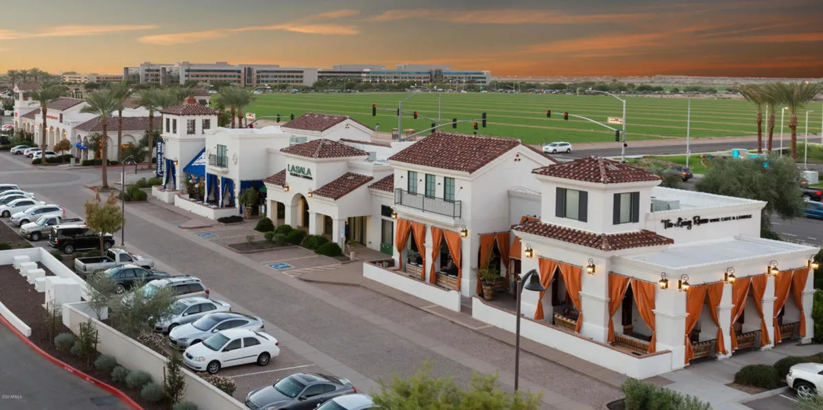 Vibrant restaurant awnings and palm trees line a lively street, with decorative architecture and a visible parking area. The atmosphere is energetic and sociable, hinting at nearby cafes and restaurants in Downtown Ocotillo.