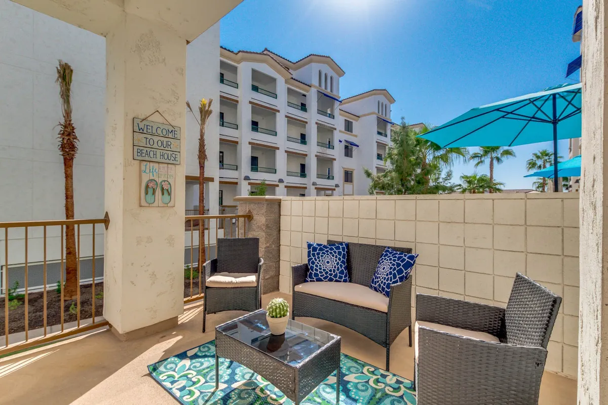 A cozy and inviting outdoor seating area with cushioned chairs, a decorative welcome sign, and vibrant pillows. The space is designed for relaxation and socializing, set against the backdrop of a well-kept patio area that suggests a connection to the surrounding greenbelt or community grounds.