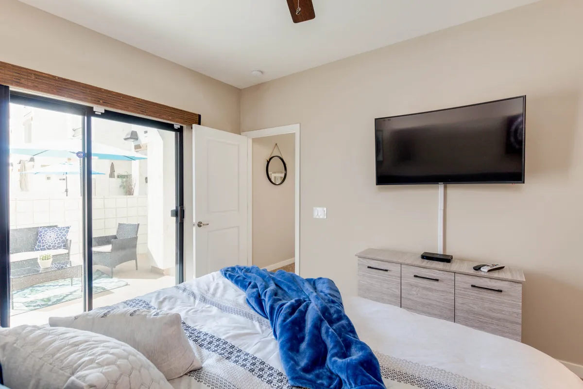 A serene bedroom with a plush bed adorned with a blue blanket. The room features a wall-mounted television, ceiling fan, and a sliding glass door that opens directly to a private outdoor patio. The space is bathed in natural light and feels tranquil and inviting.