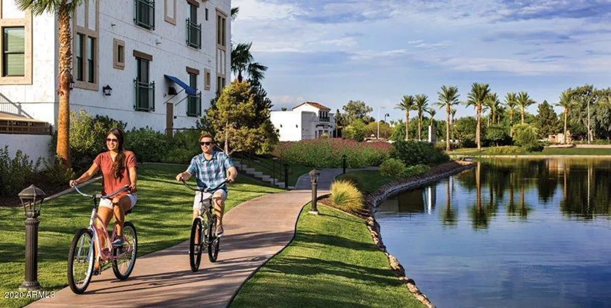 An exterior scene featuring bicyclists riding along a paved lakeside pathway lined with palm trees, with modern buildings and a serene lake in the background. The atmosphere is vibrant and active, suggesting a lively community and opportunities for outdoor activities.