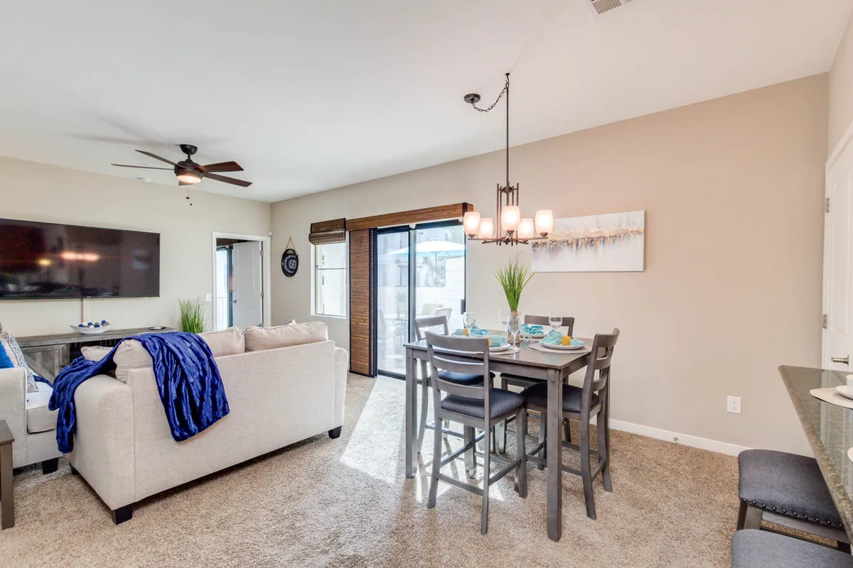 A stylish open-concept great room featuring a modern sofa, a full dining table under a contemporary chandelier, and a large TV mounted on the wall. The seamless layout visually unites the living, dining, and lounge areas, creating a welcoming space ideal for relaxing or entertaining.