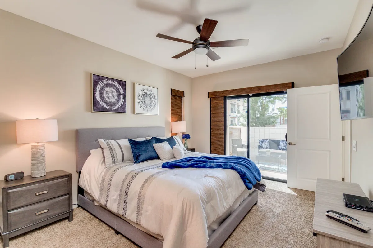 A bright, serene bedroom with a bed draped in a blue throw blanket and a large window offering an inviting view outside. There is a contemporary nightstand with a lamp and tasteful artwork on the wall, evoking a calm, romantic mood.